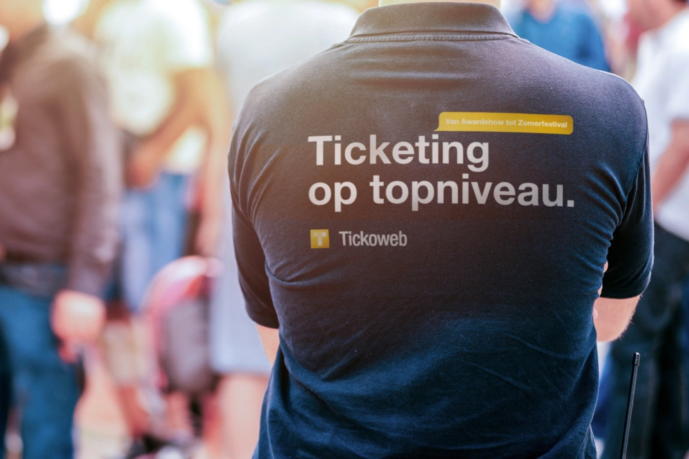 De achterzijde van het T-shirt van een Tickoweb medewerker. De slogan op het T-shirt luidt "Ticketing op topniveau".